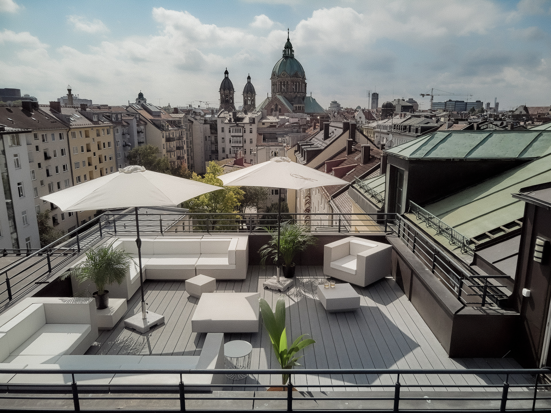 Modern rooftop terrace with white furniture, umbrellas, plants, overlooking city buildings and domed cathedral. Showing the Tattoo Studio Pardon Paris in munich