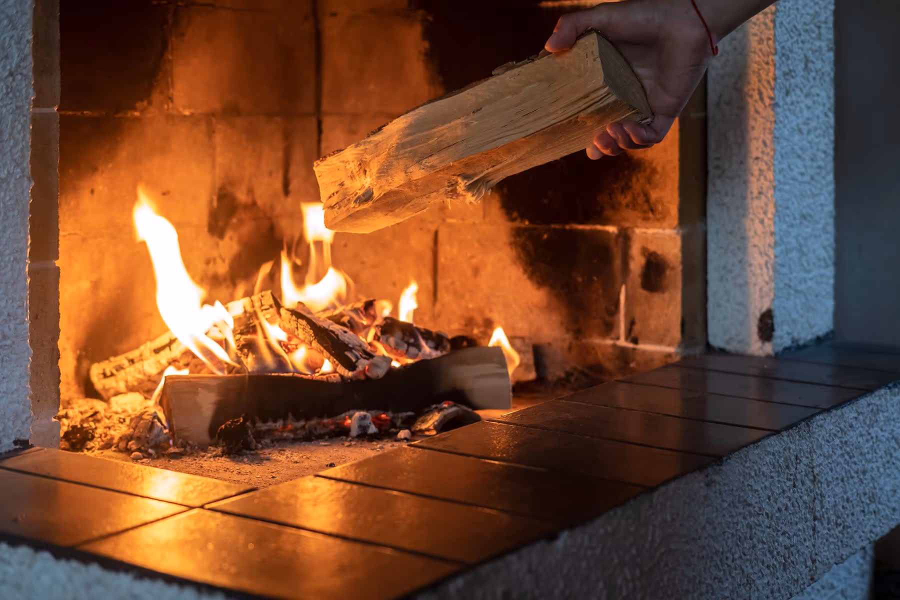 En hånd legger vedkubber i en peis med levende flammer.