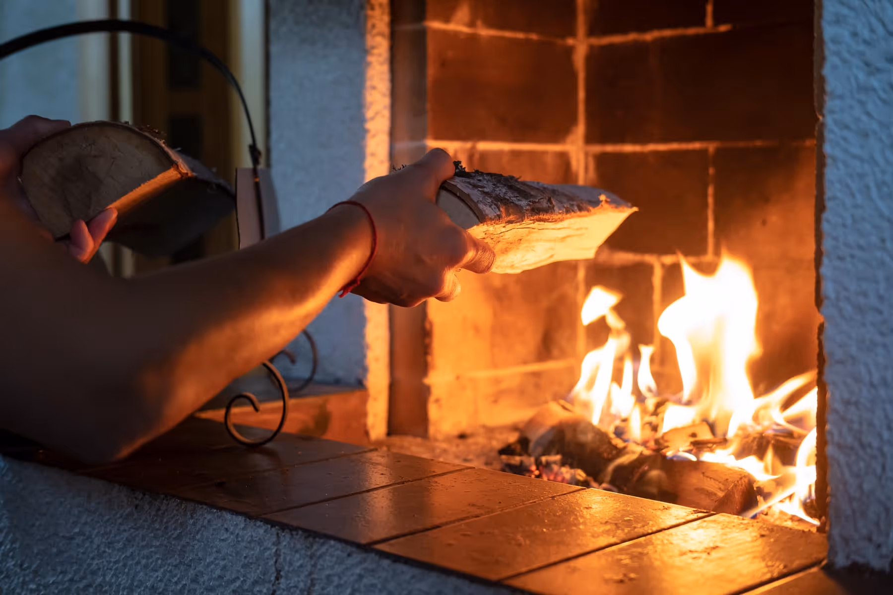 Hender legger til vedkubber i en åpen peis med flammer som lyser opp området.