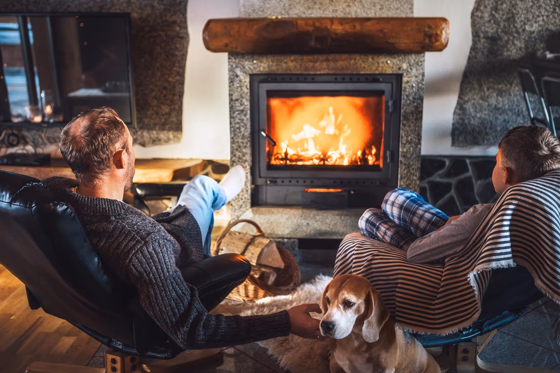 To mennisker sitter ved en peis med en hund i en koselig stue.