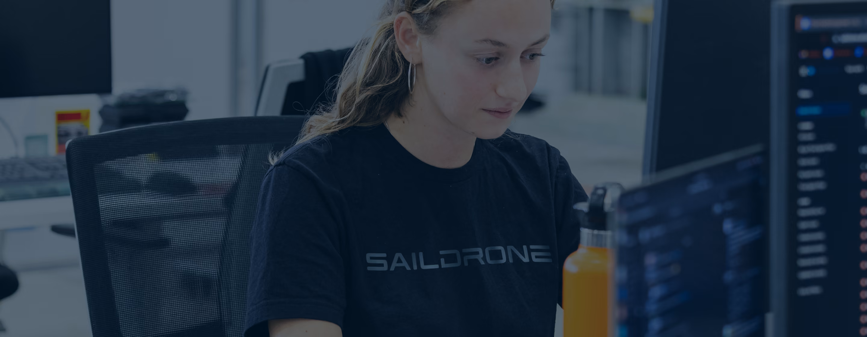 Woman wearing a Saildrone shirt working at a computer with an orange water bottle nearby.