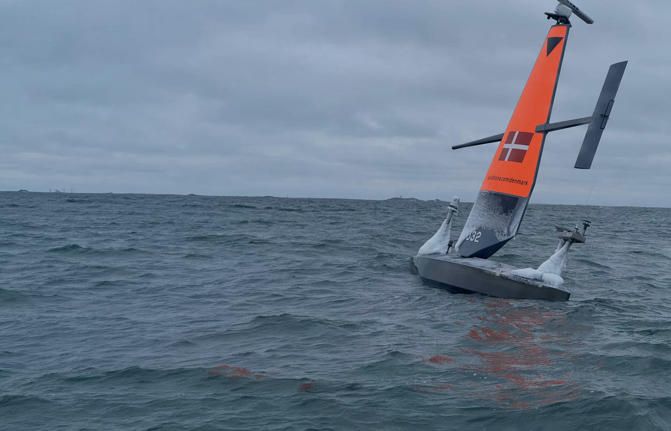 Autonomous Saildrone with an orange sail and Danish flag floating on choppy ocean water under gray sky.