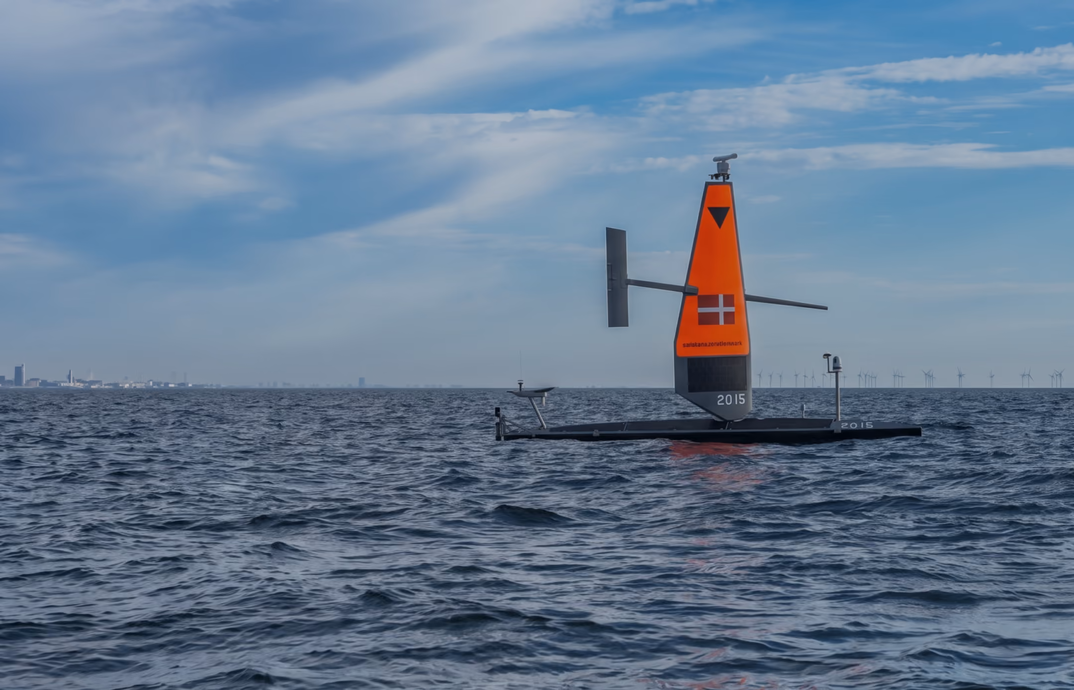 Saildrone with Danish flag floating on the ocean with city skyline and wind turbines in the background.