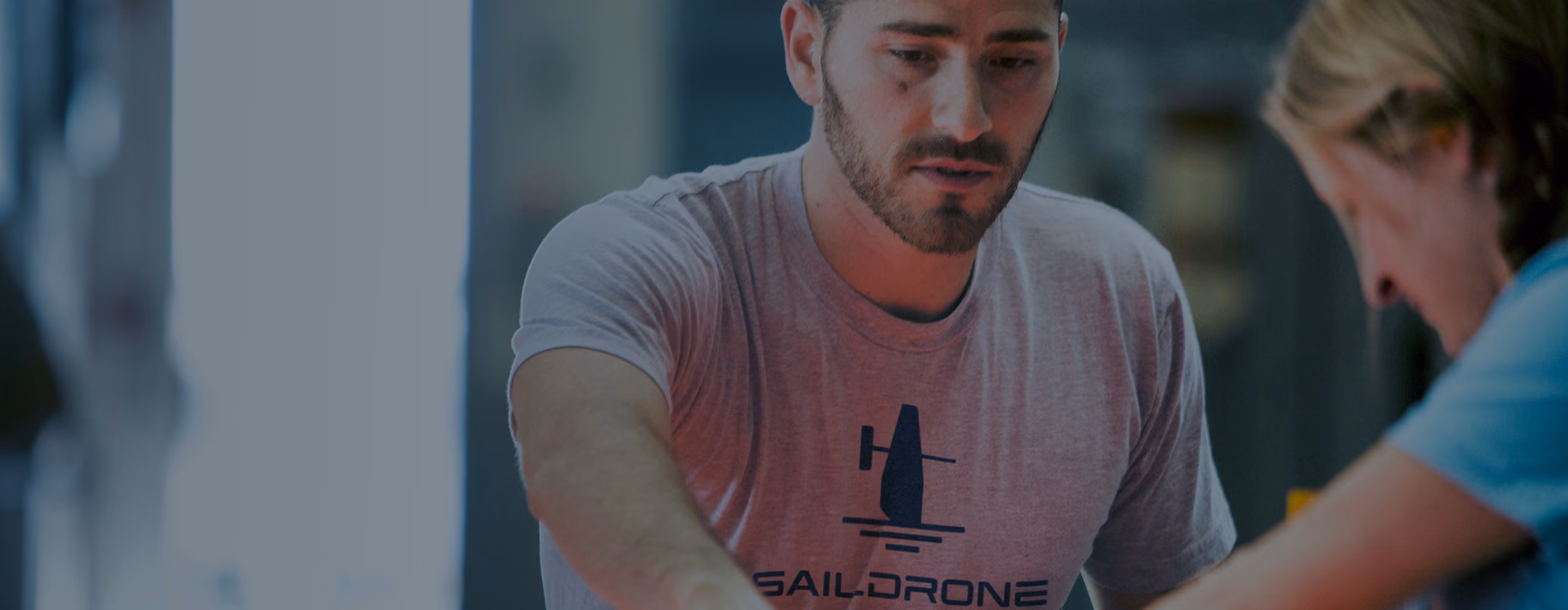 Man with beard in a Saildrone t-shirt engaged in a focused activity indoors with another person.