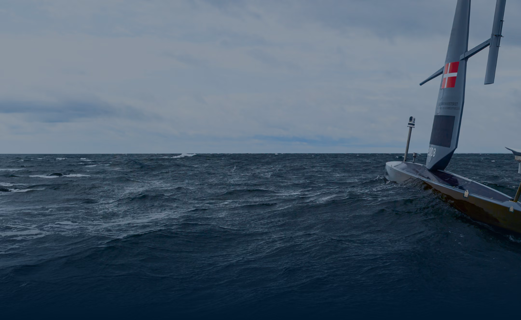 Saildrone with a Danish flag on a dark blue ocean under a cloudy sky.