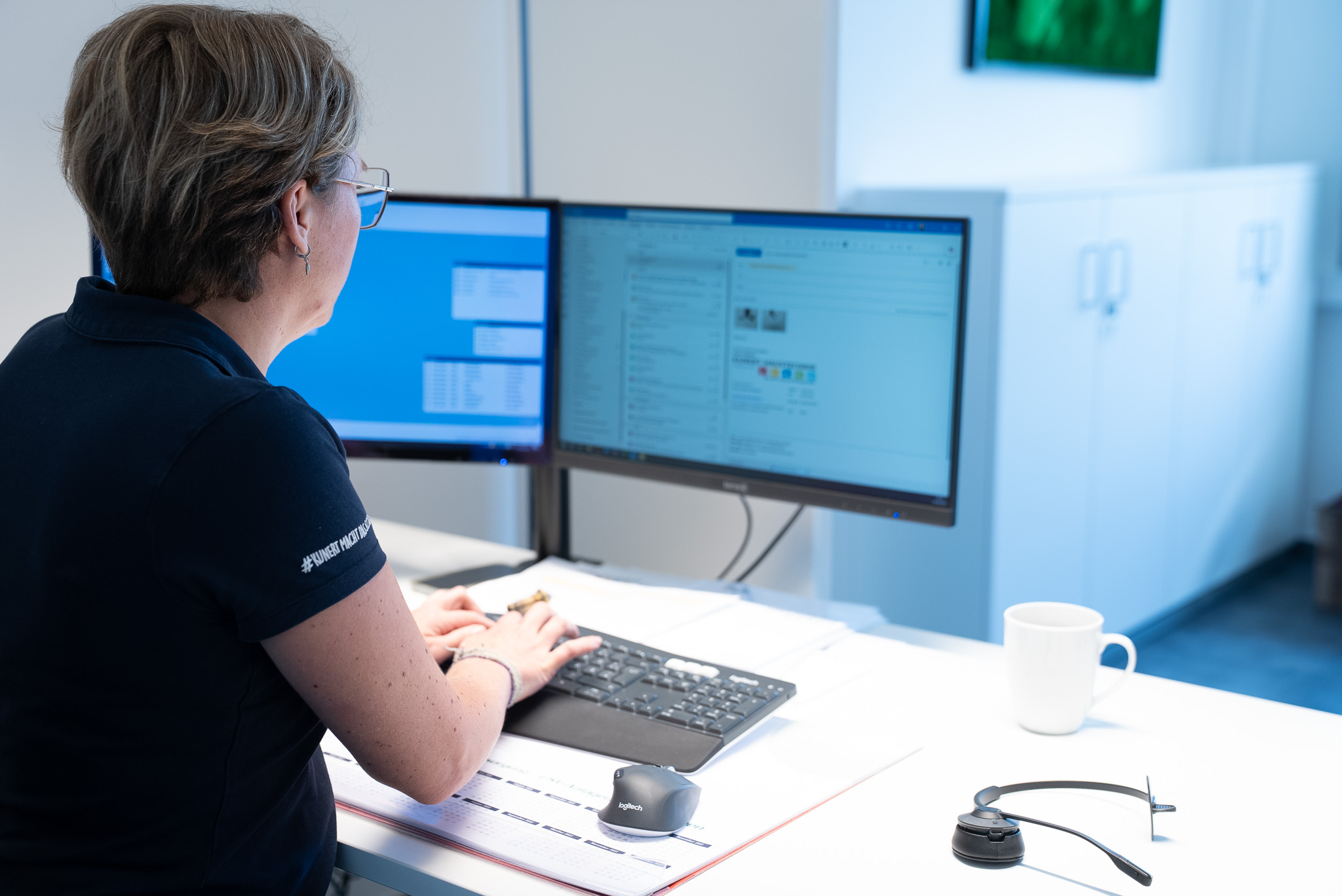 Frau arbeitet an einem Schreibtisch mit zwei Monitoren, Tastatur, Maus und Headset.
