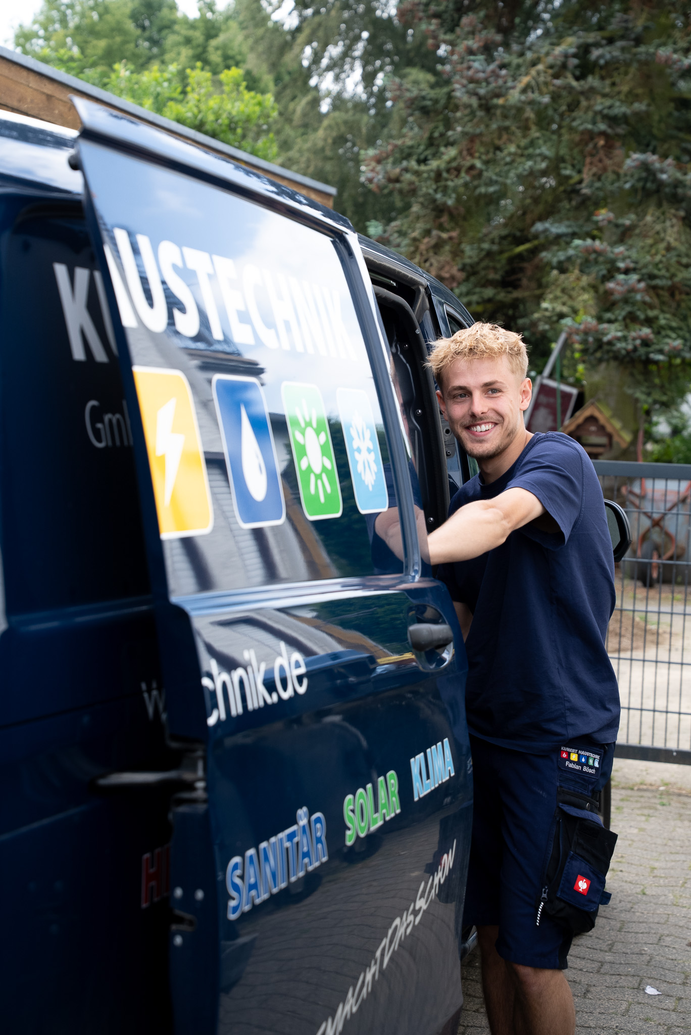 Junger Handwerker lächelt neben einem blauen Firmenfahrzeug mit Sanitär-, Solar- und Klimadienstleistungen.