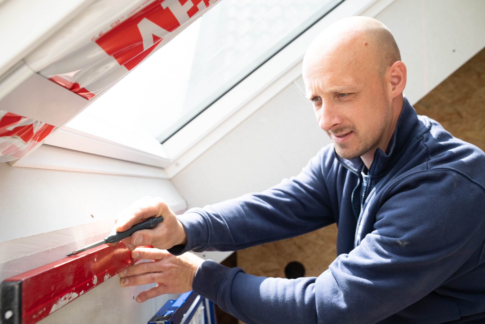 Mann befestigt mit Schraubendreher ein rotes Werkzeug an einer Wand unter einem Dachfenster.