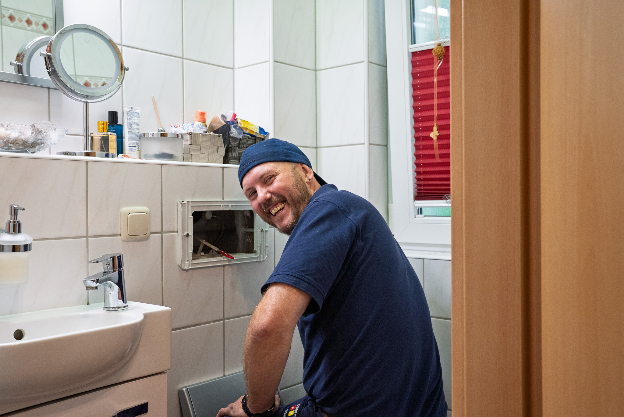 Mann in blauer Kleidung repariert Waschbecken im hellen Badezimmer und lächelt zur Kamera.