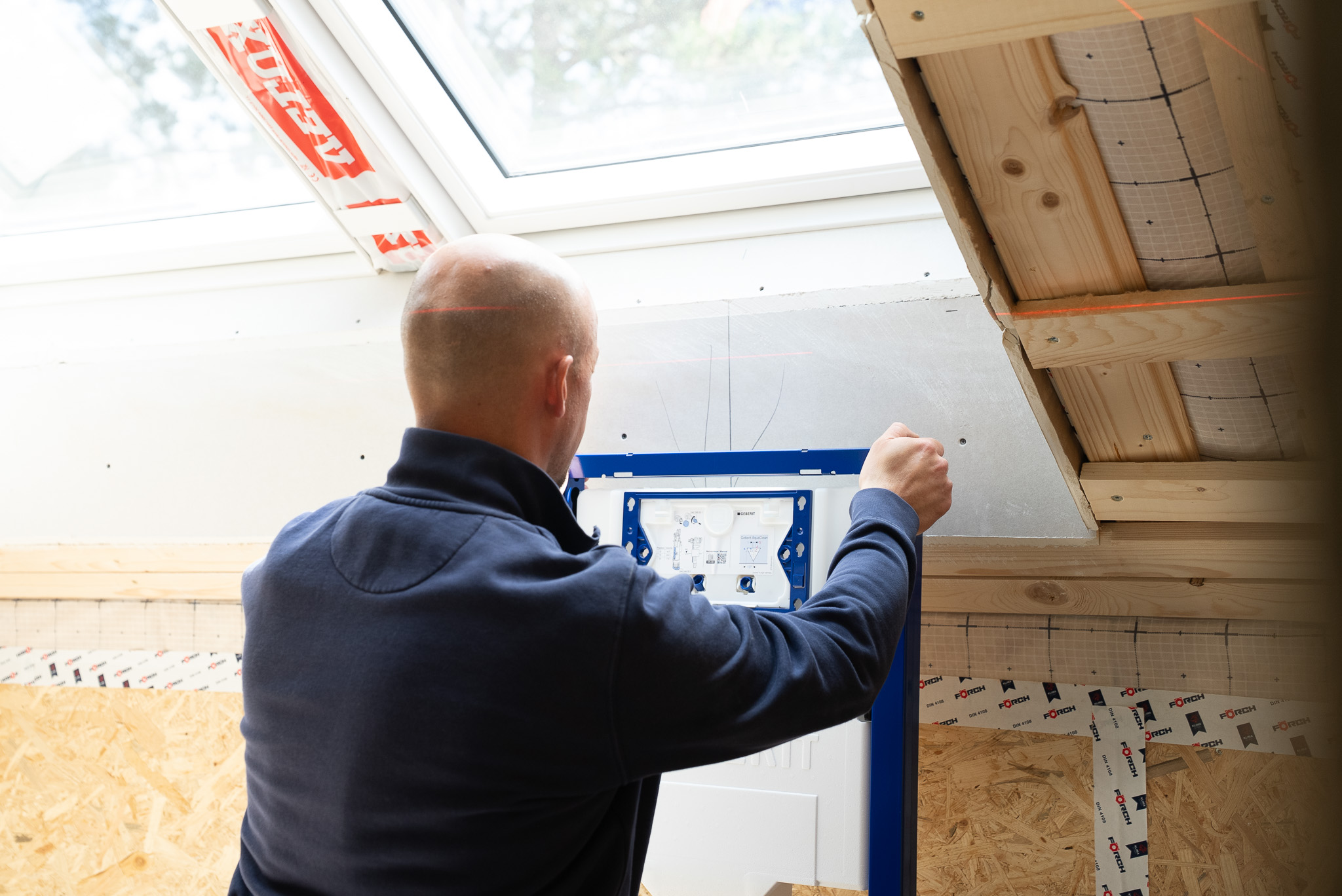 Handwerker installiert eine blaue Sanitärrahmenstruktur unter einem Dachfenster in einem unfertigen Raum.