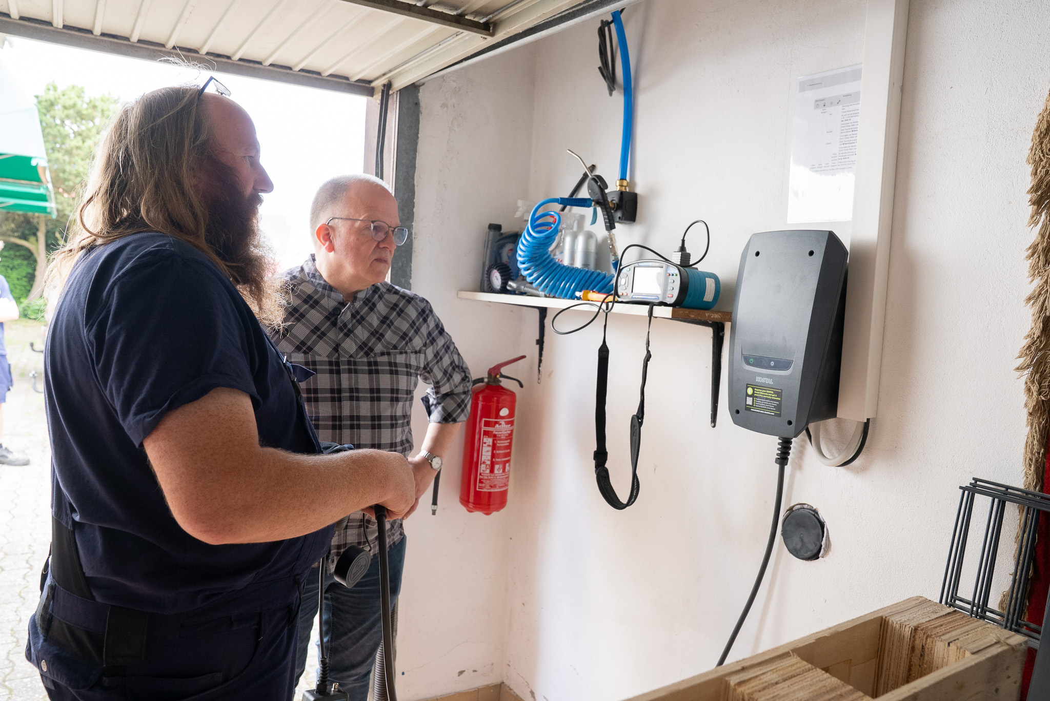 Zwei Männer prüfen technische Geräte an einer Wand in einer Garage oder Werkstatt.