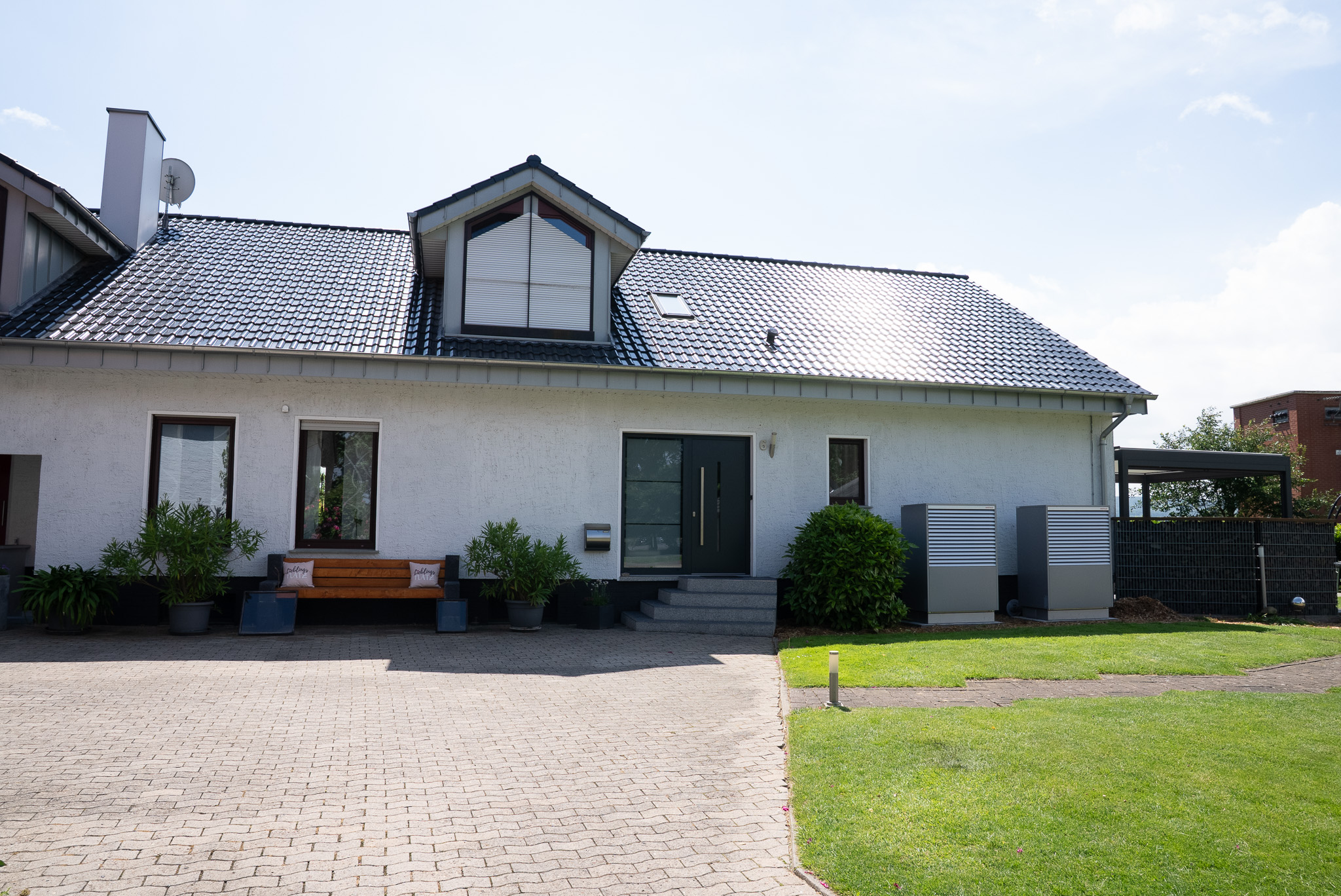 Haus mit dunklem Dach, grünem Eingang, Sitzbank, Pflanzen und gepflegtem Vorgarten bei Sonnenschein.
