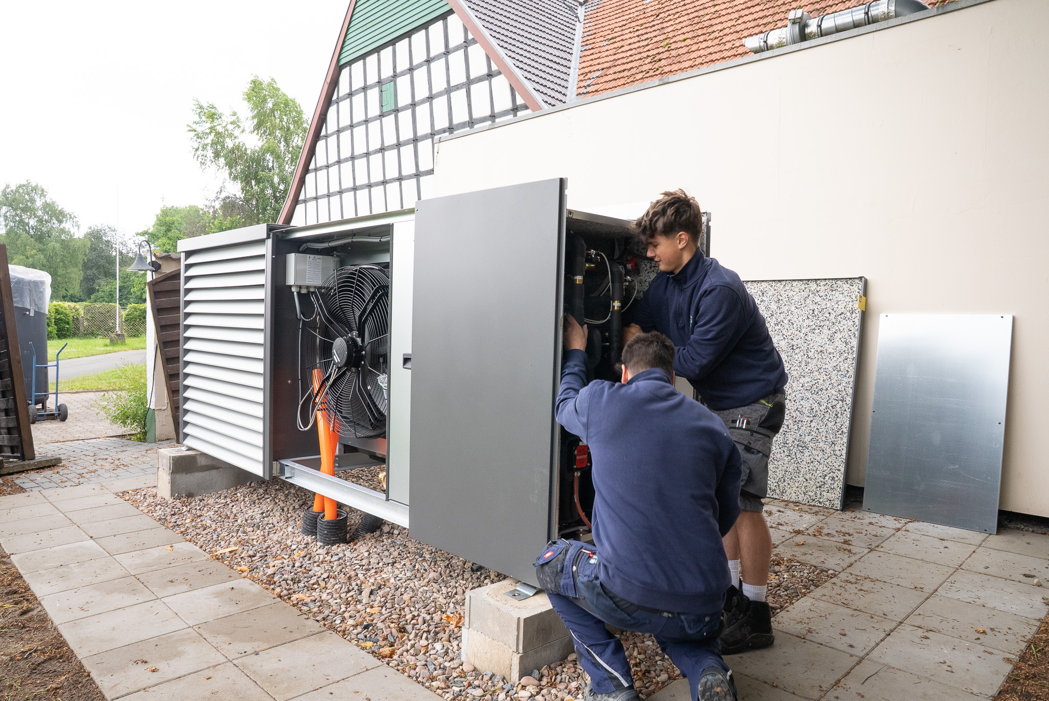 Zwei Techniker reparieren eine große Außeneinheit einer Klimaanlage oder Wärmepumpe neben einem Haus.
