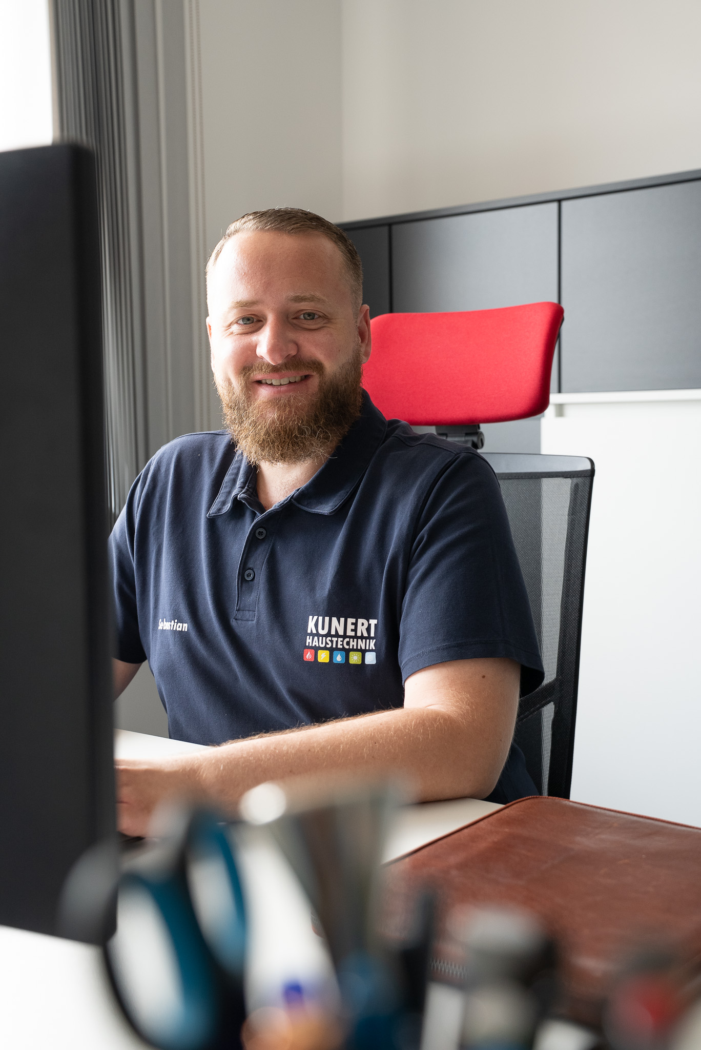 Lächelnder Mann im dunkelblauen Kunert Haustechnik-Poloshirt sitzt am Schreibtisch vor einem Computer.