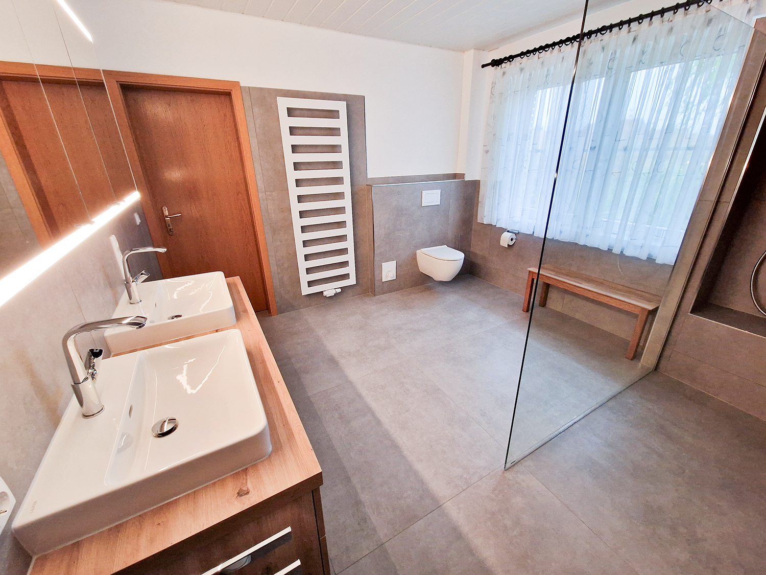 Modernes Badezimmer mit Doppelwaschbecken, wandhängendem WC, Bank und großer Dusche mit Glaswand.