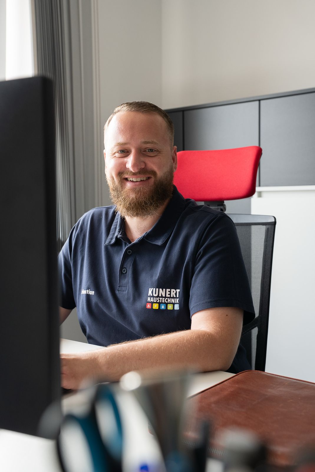 Lächelnder Mann mit Bart sitzt am Schreibtisch in einem Büro und trägt ein Polohemd mit Firmenlogo.