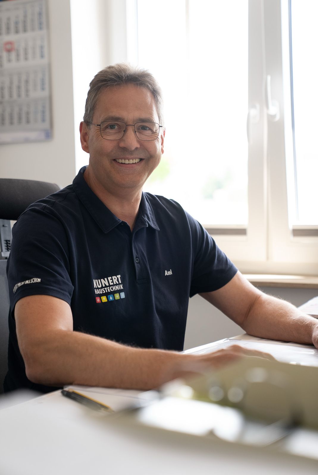 Lächelnder Mann mit Brille trägt ein Kunert Haustechnik Polohemd mit Namen Axel im Büro.