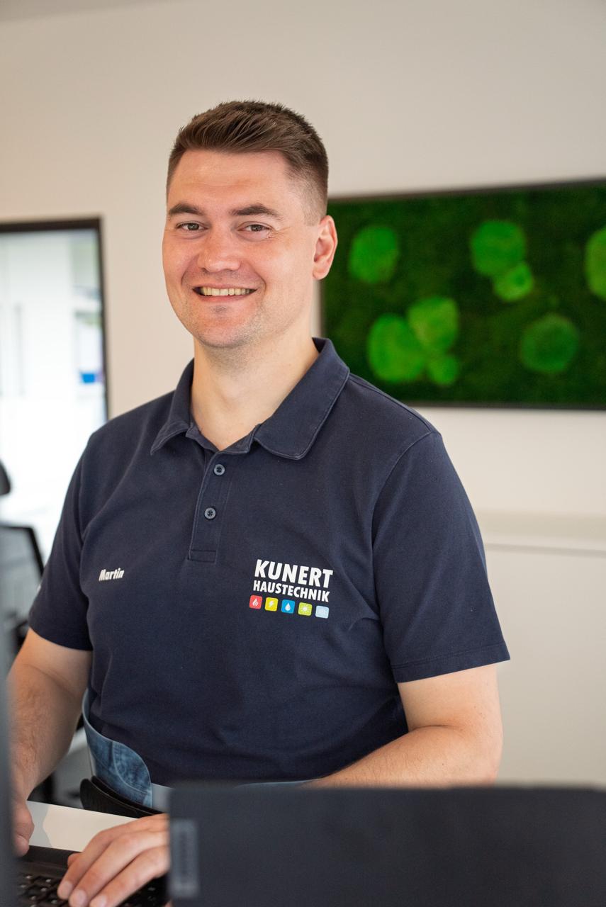 Smiling man wearing a navy Kunert Haustechnik shirt sitting at a desk with a laptop.