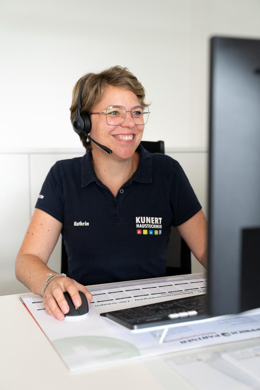 Frau mit Brille und Headset lächelt am Schreibtisch vor Computer, trägt Kunert Haustechnik Polo.