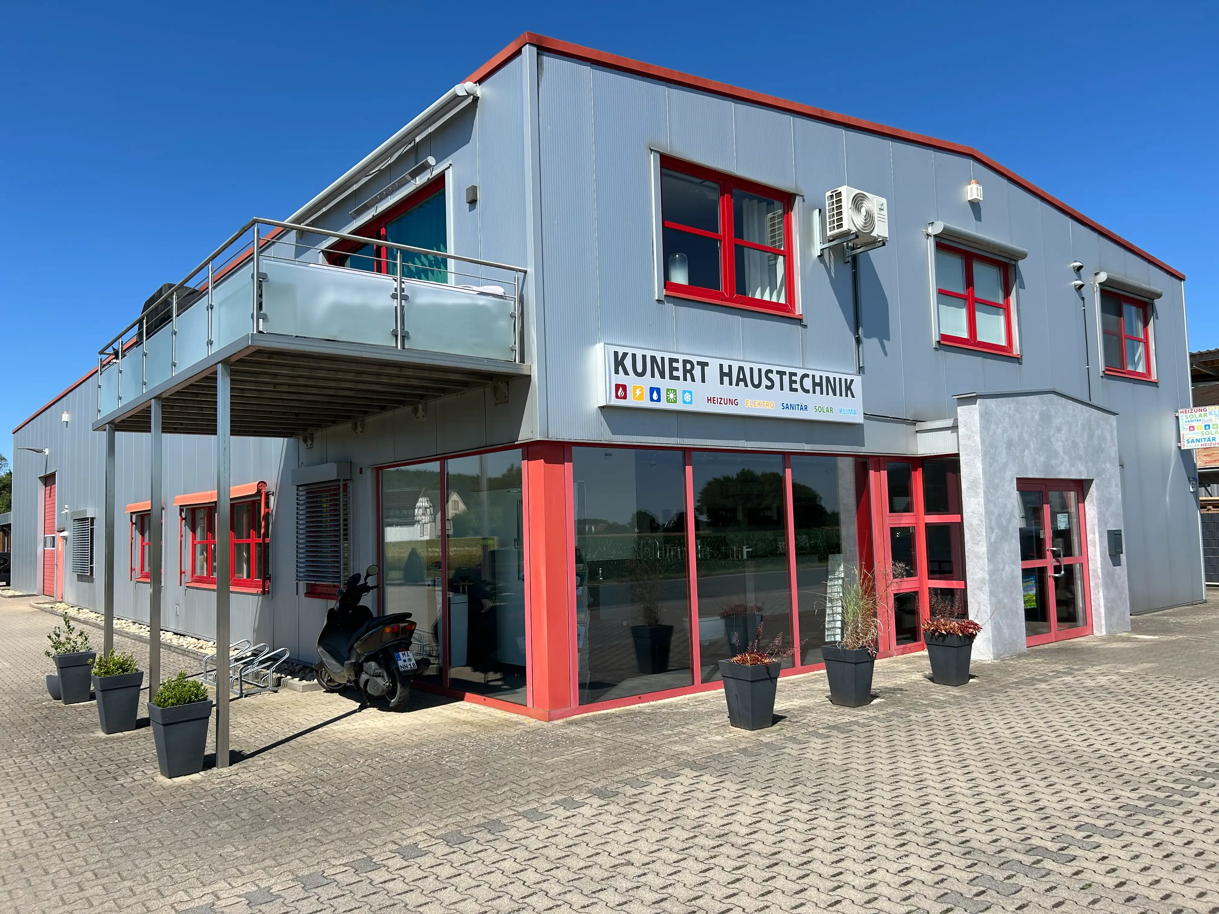 Modernes graues Gebäude mit roten Fensterrahmen und Schild Kunert Haustechnik unter blauem Himmel.