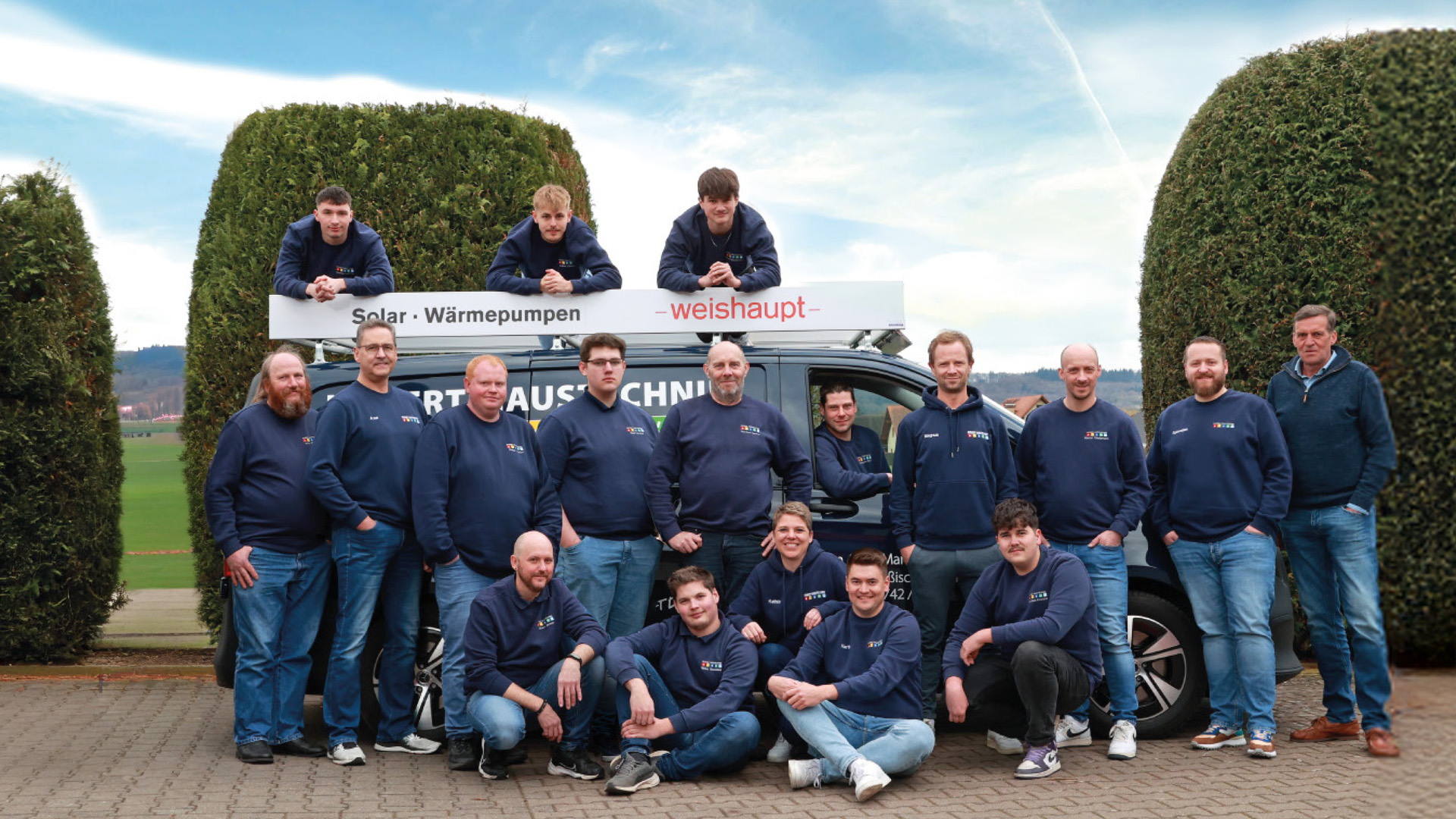 Gruppenfoto von Teammitgliedern in blauen Pullovern vor einem Fahrzeug mit Solar- und Wärmepumpenwerbung.