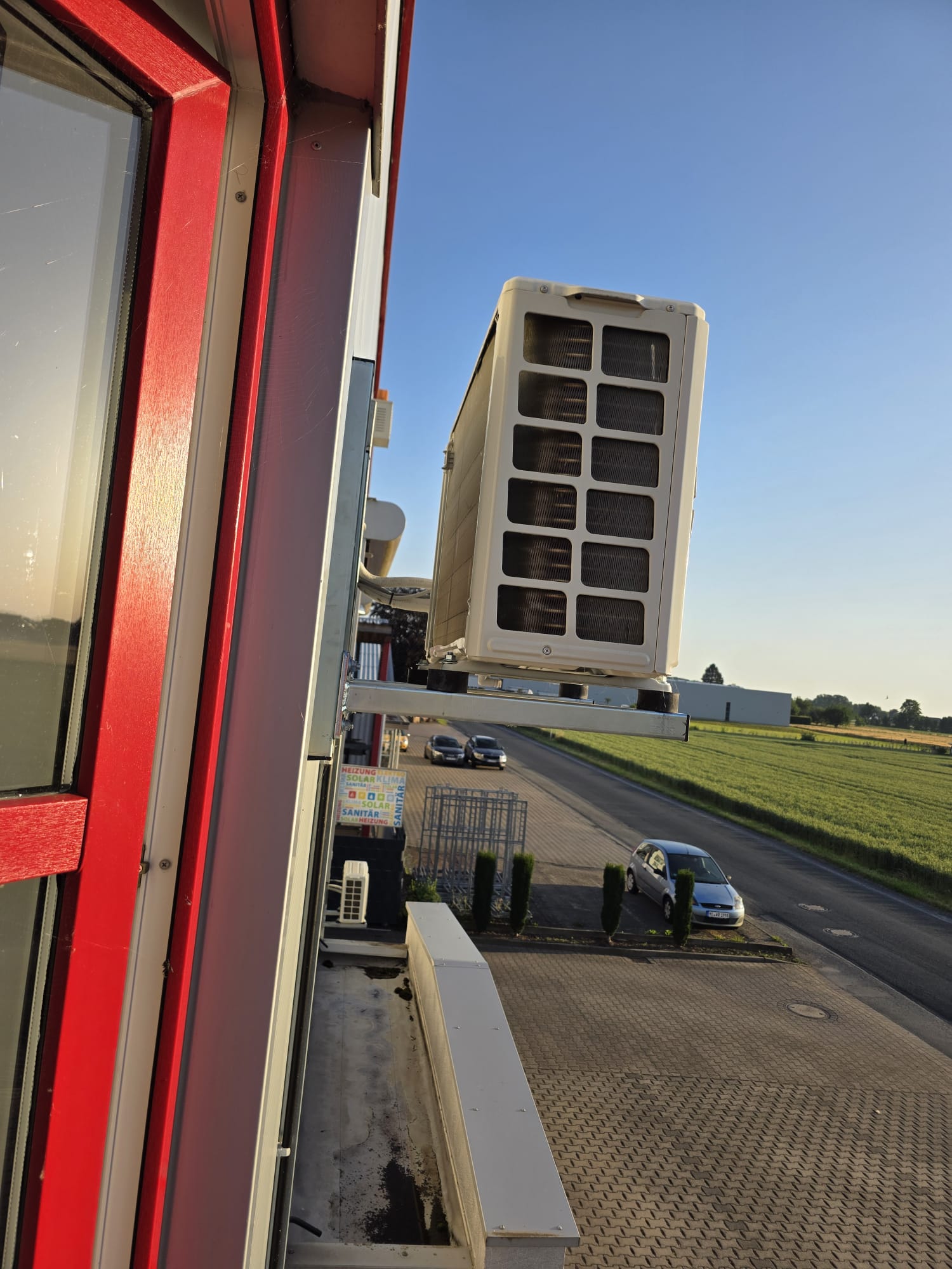 Außenklimaanlage an Gebäudewand mit Parkplatz und Wiese im Hintergrund bei klarem Himmel.