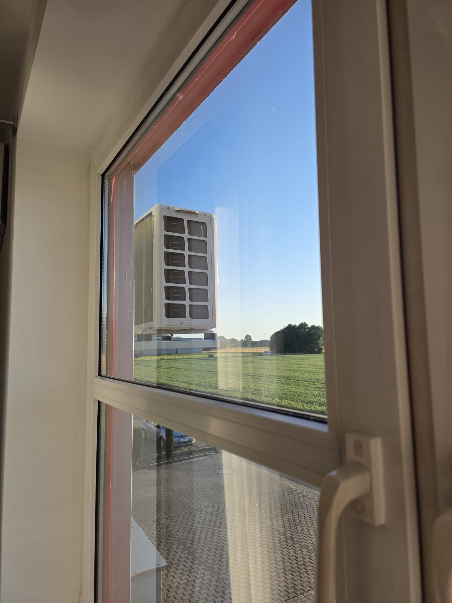 Blick aus einem Fenster auf eine Klimaanlage mit grüner Wiese und blauem Himmel im Hintergrund.