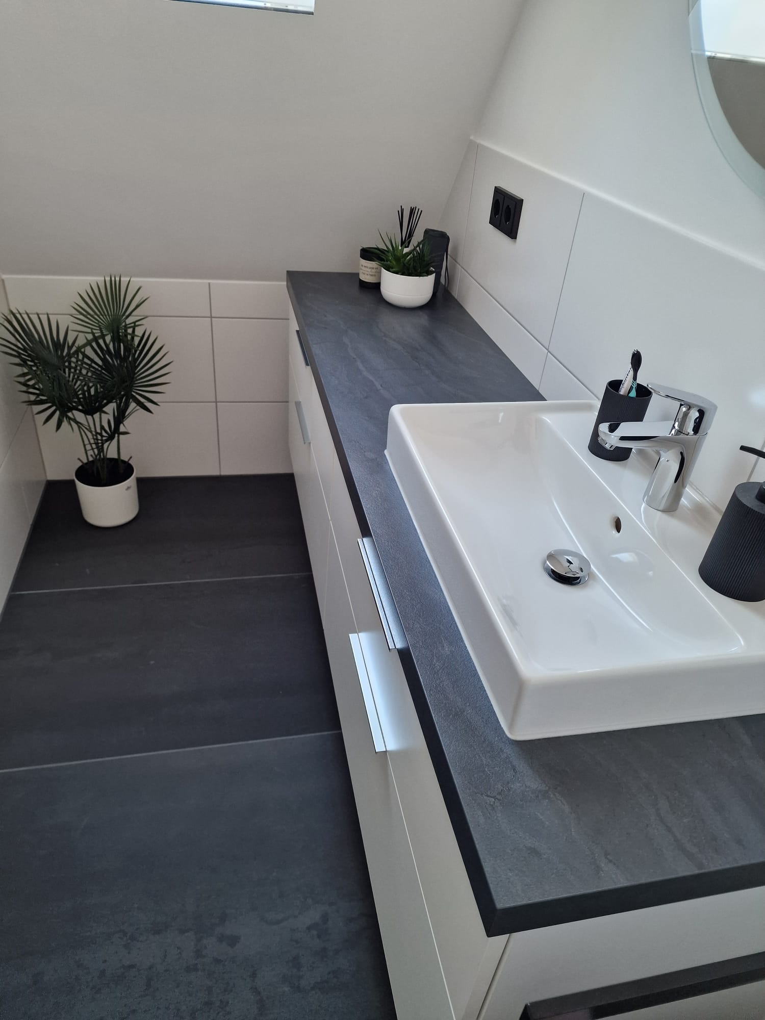 Modernes Badezimmer mit weißem Waschbecken, dunkler Arbeitsplatte und grünen Pflanzen in Töpfen.