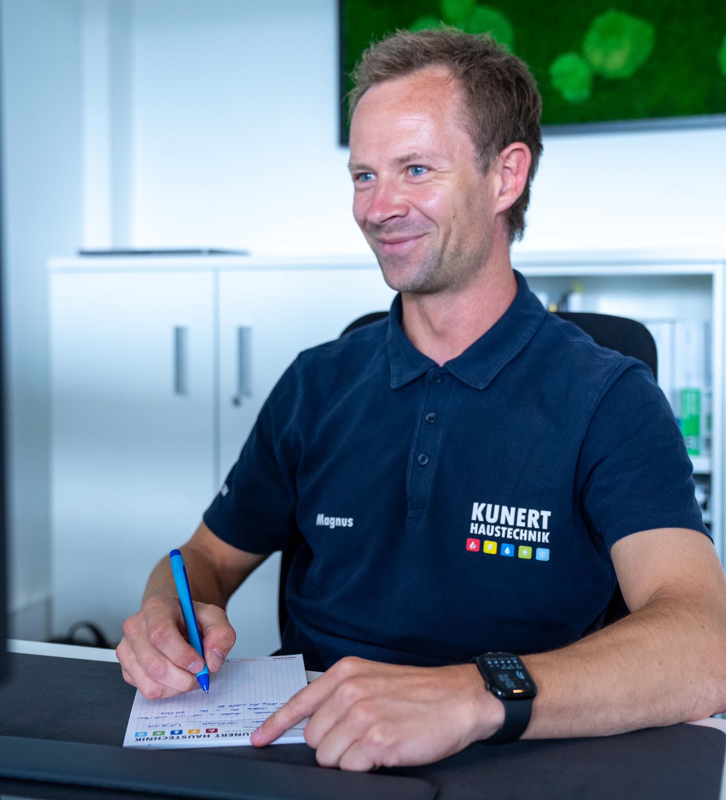 Mann in blauem Kunert Haustechnik Poloshirt sitzt lächelnd am Schreibtisch und schreibt auf Papier.