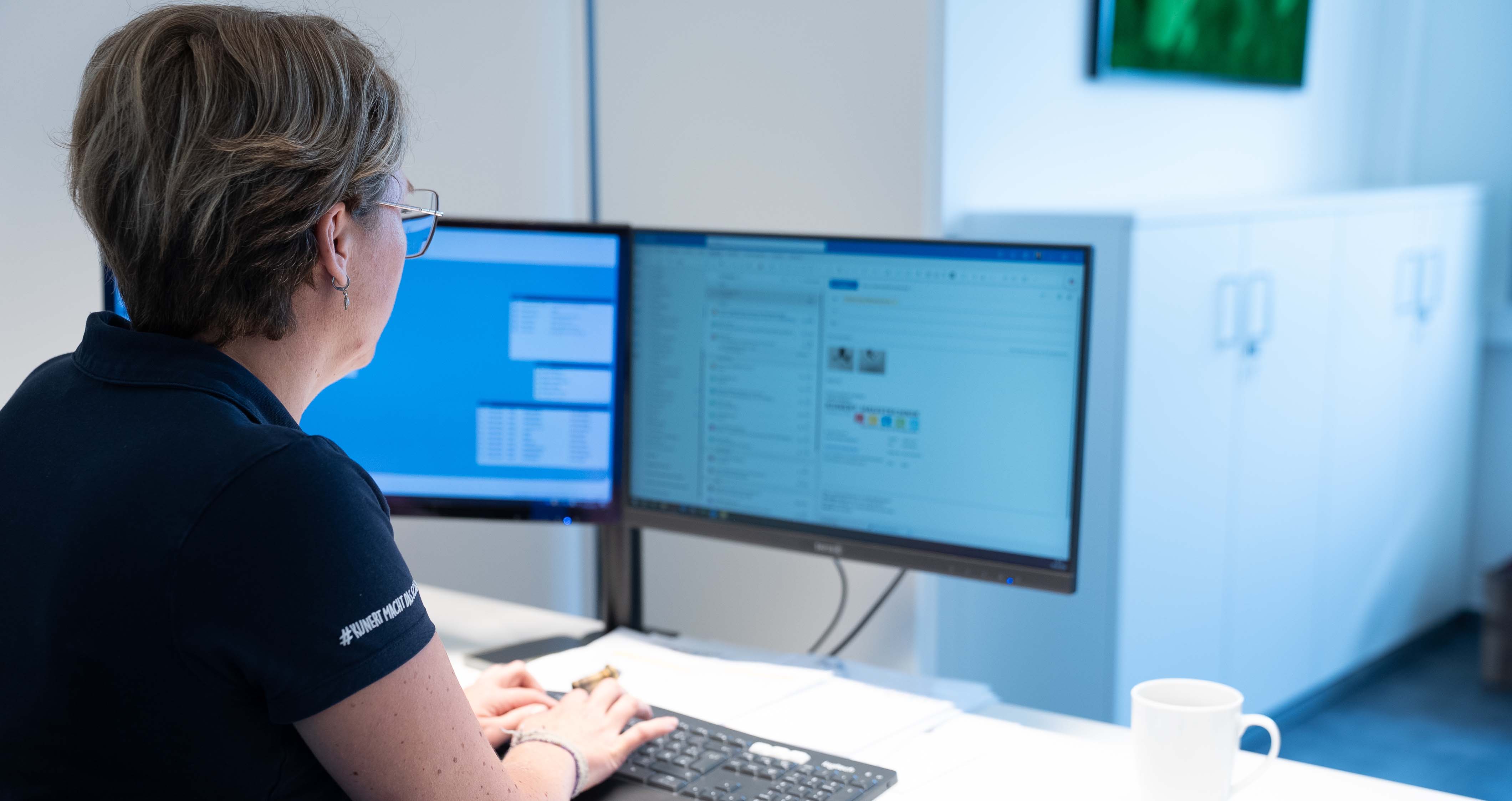 Frau arbeitet an einem Schreibtisch mit zwei Computermonitoren und Tastatur in hellem Büro.