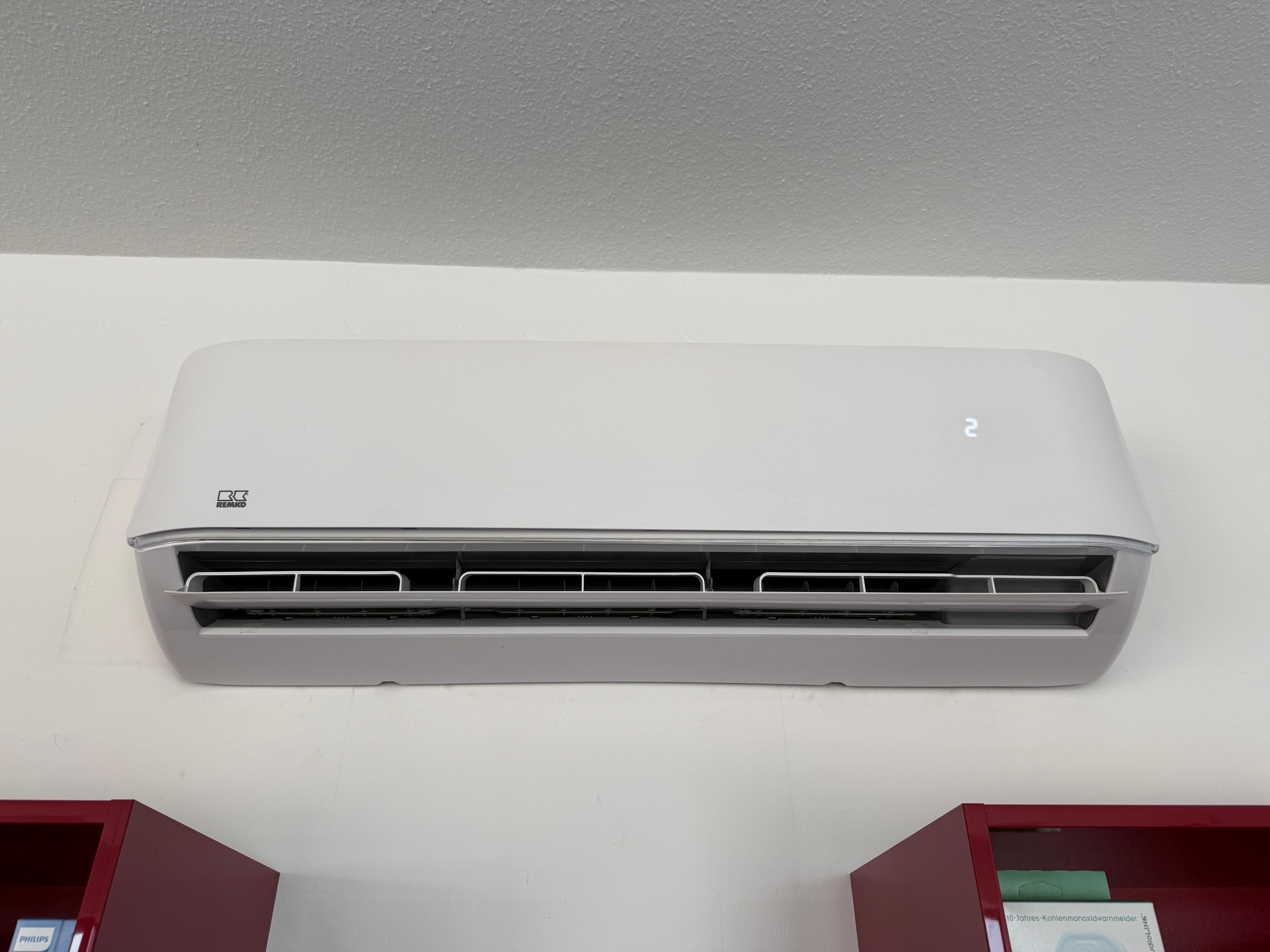Weiße Wand-Klimaanlage mit offenem Auslass und digitaler Anzeige über roten Regalen.