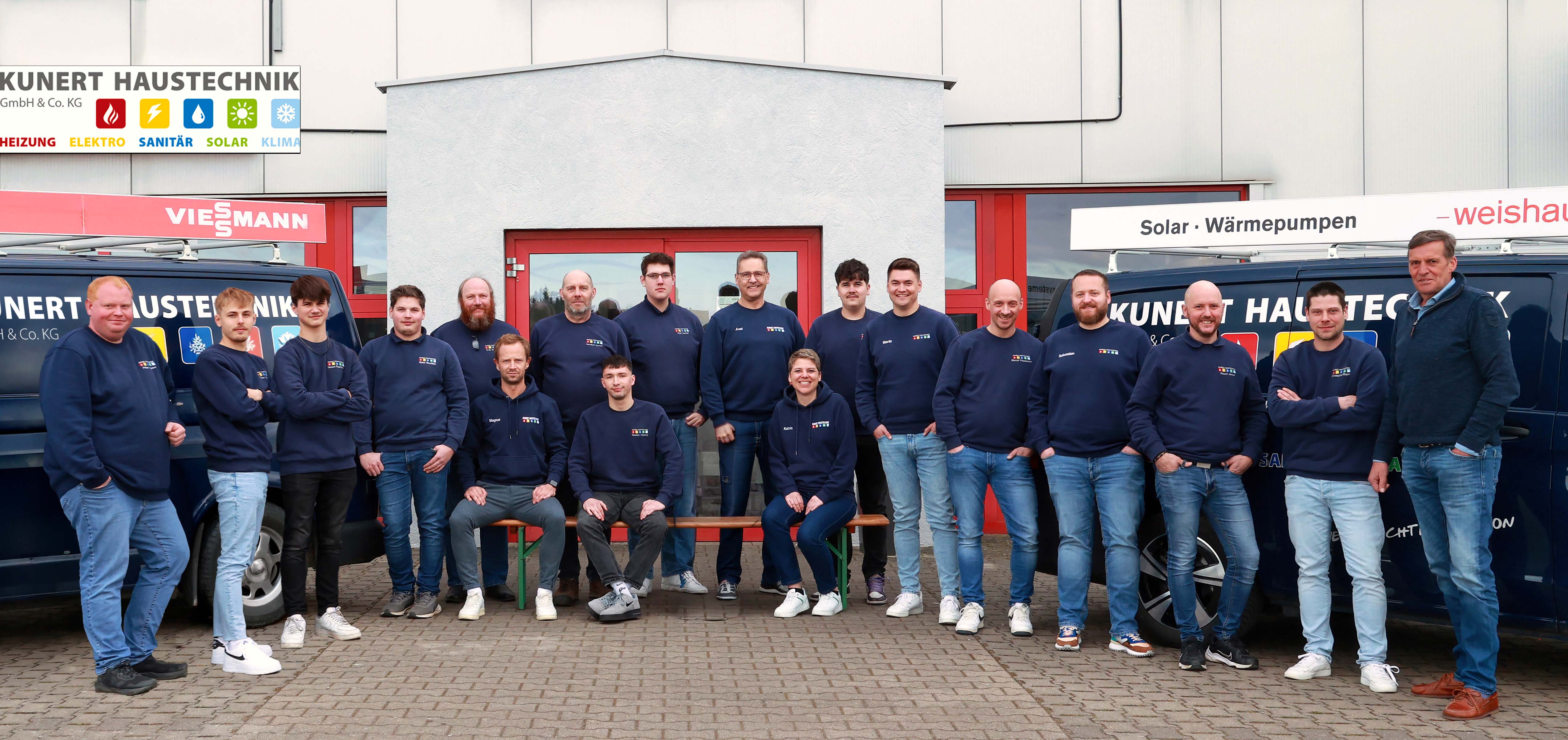 Team von 16 Personen in blauen Sweatshirts und Jeans vor zwei Firmenfahrzeugen mit Firmenlogo.