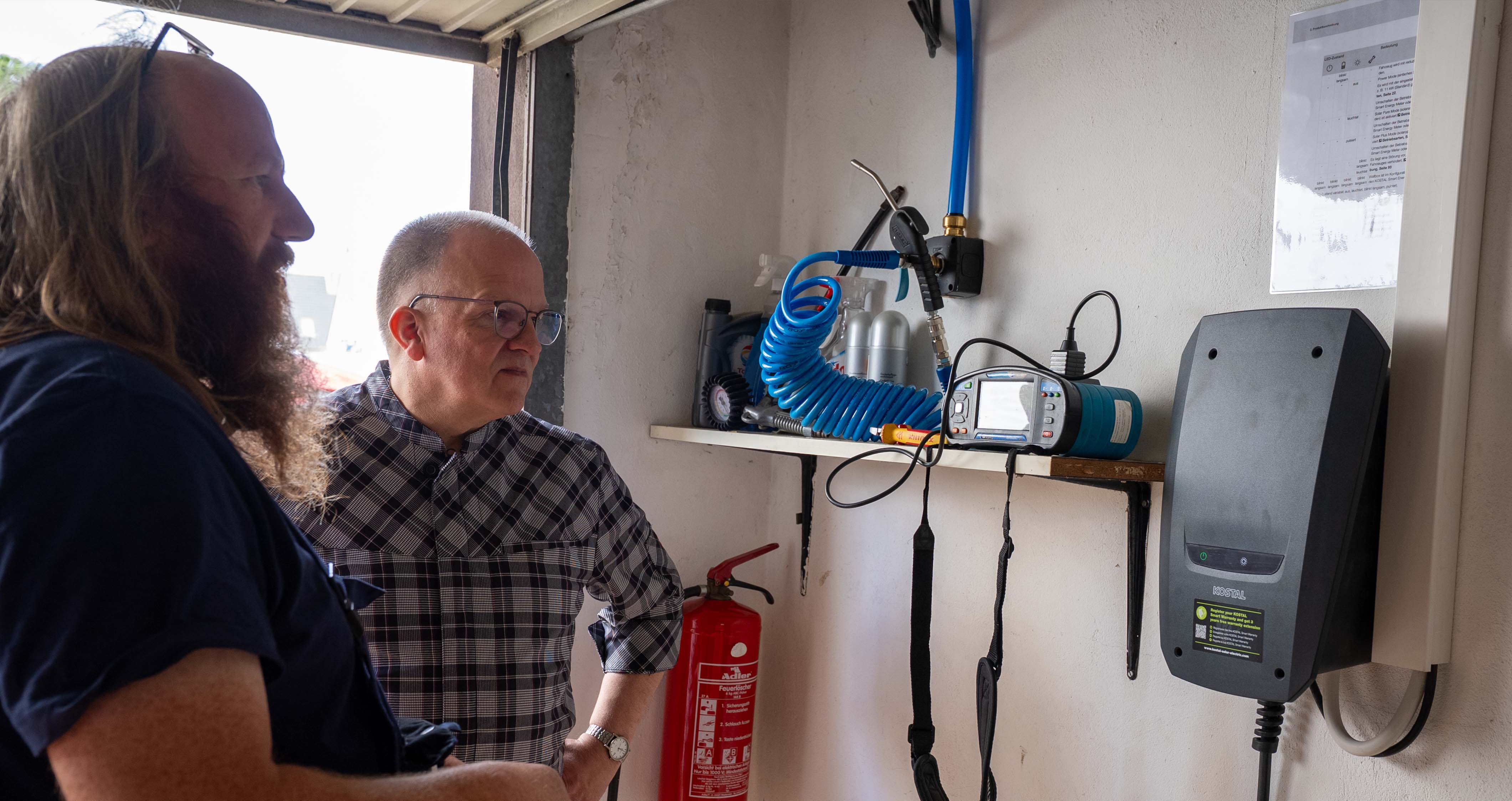 Zwei Männer betrachten ein Wandgerät mit blauer Spiralschlaucheinheit und einem Feuerlöscher daneben.
