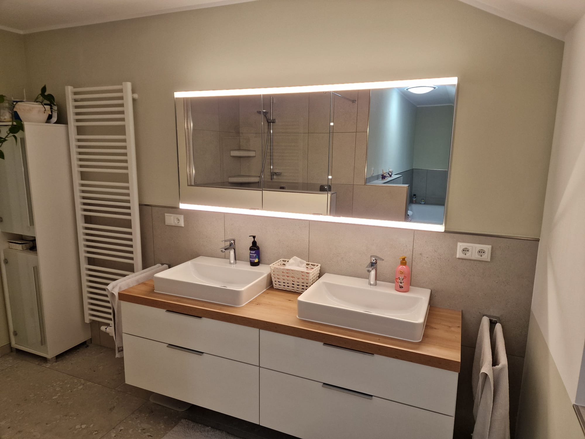 Modernes Badezimmer mit zwei weißen Waschbecken auf Holzplatte und beleuchtetem Wandspiegel.