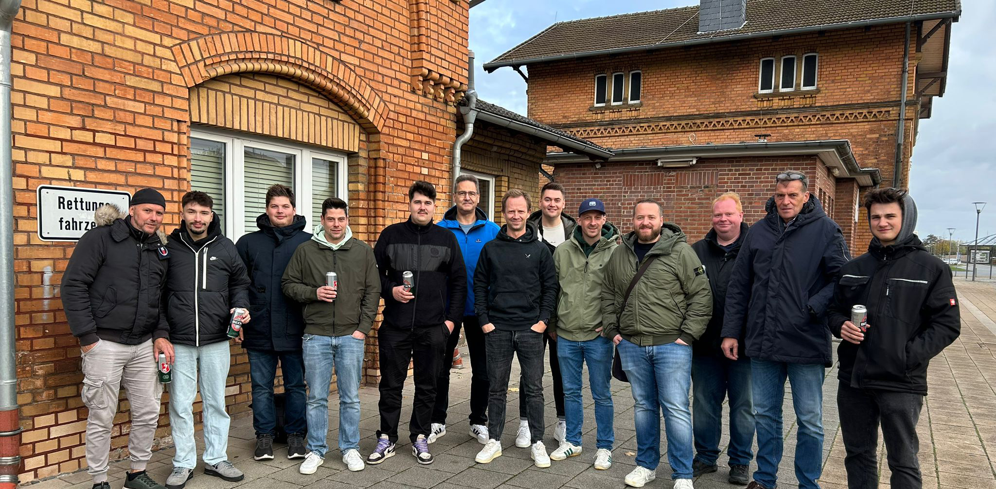 Gruppenfoto von 14 Männern vor roten Backsteingebäuden, einige halten Getränkedosen.