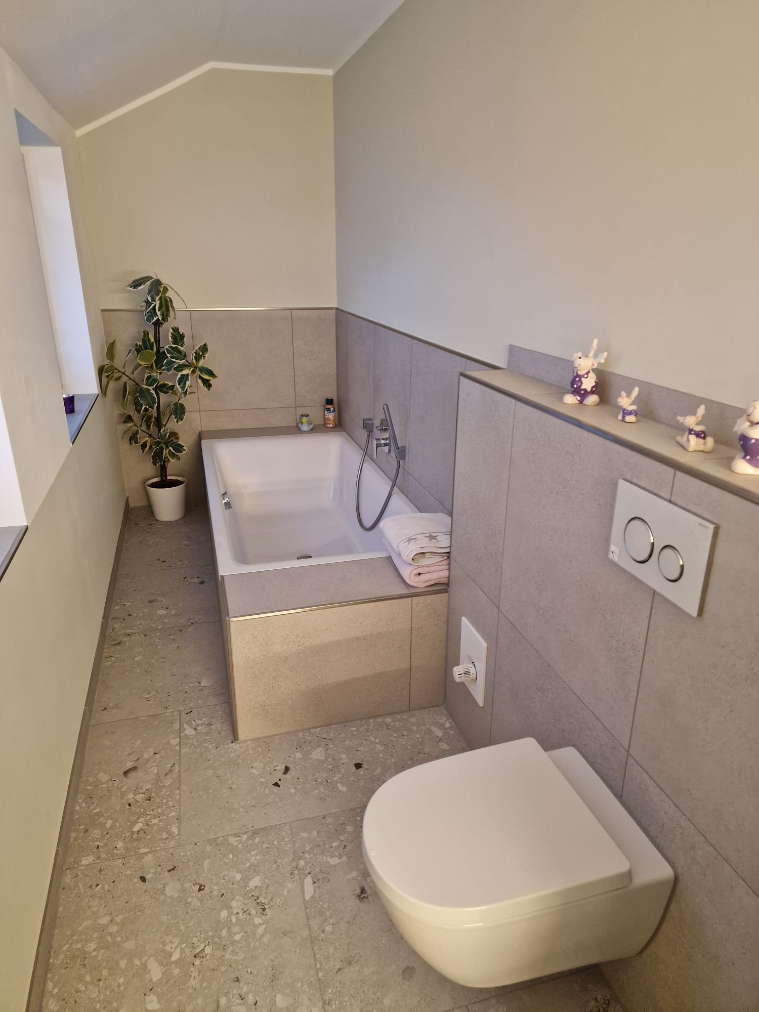Modernes Badezimmer mit eingebauter Badewanne, Wandtoilette, Pflanzen und Dekofiguren.