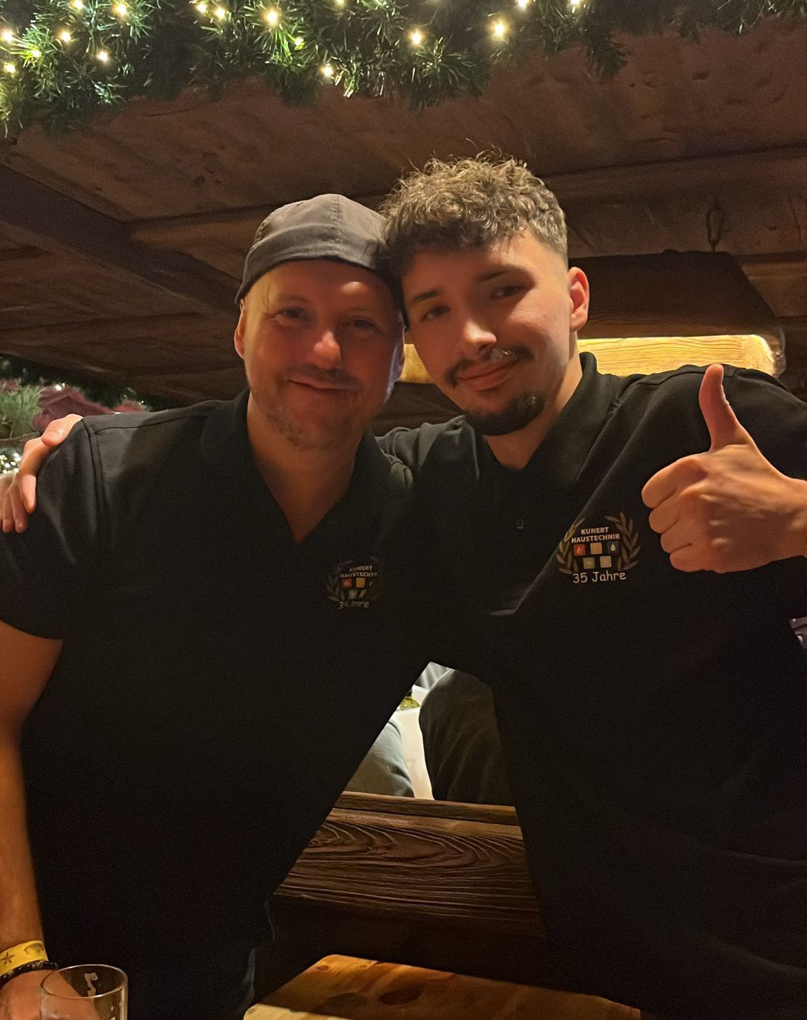 Zwei Männer in schwarzen Polo-Shirts posieren mit Arm um Schulter, einer zeigt Daumen hoch.