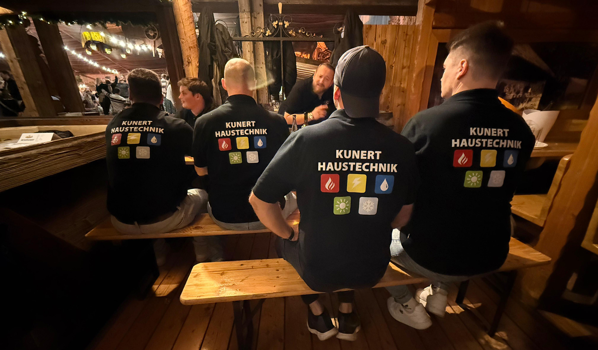 Vier Männer sitzen auf Holzbank, tragen schwarze T-Shirts mit dem Aufdruck Kunert Haustechnik und Symbolen.