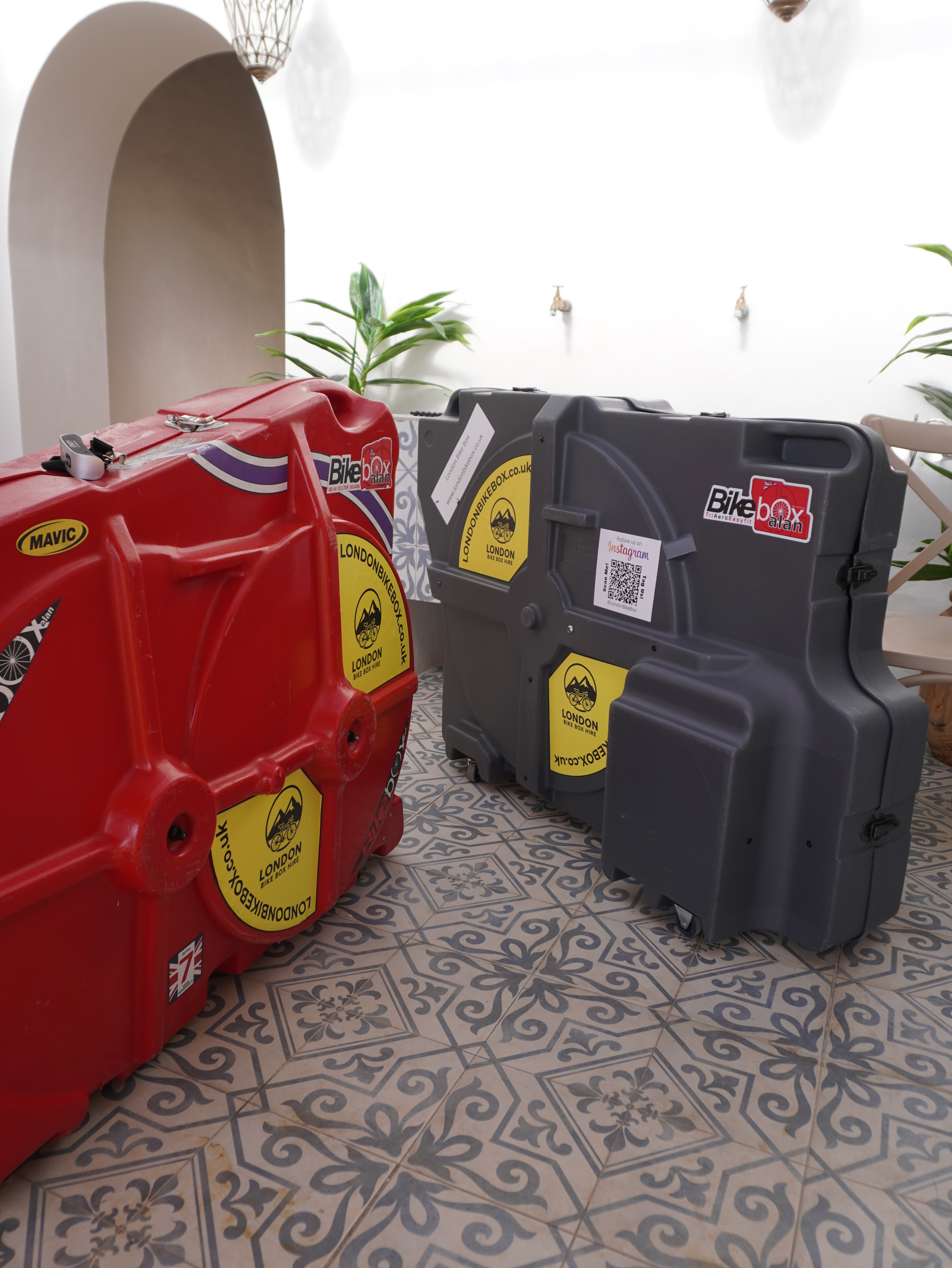 Red and gray hard bike transport cases with LondonBikeBox.co.uk stickers placed on patterned tile floor inside a bright room with arch and plants.
