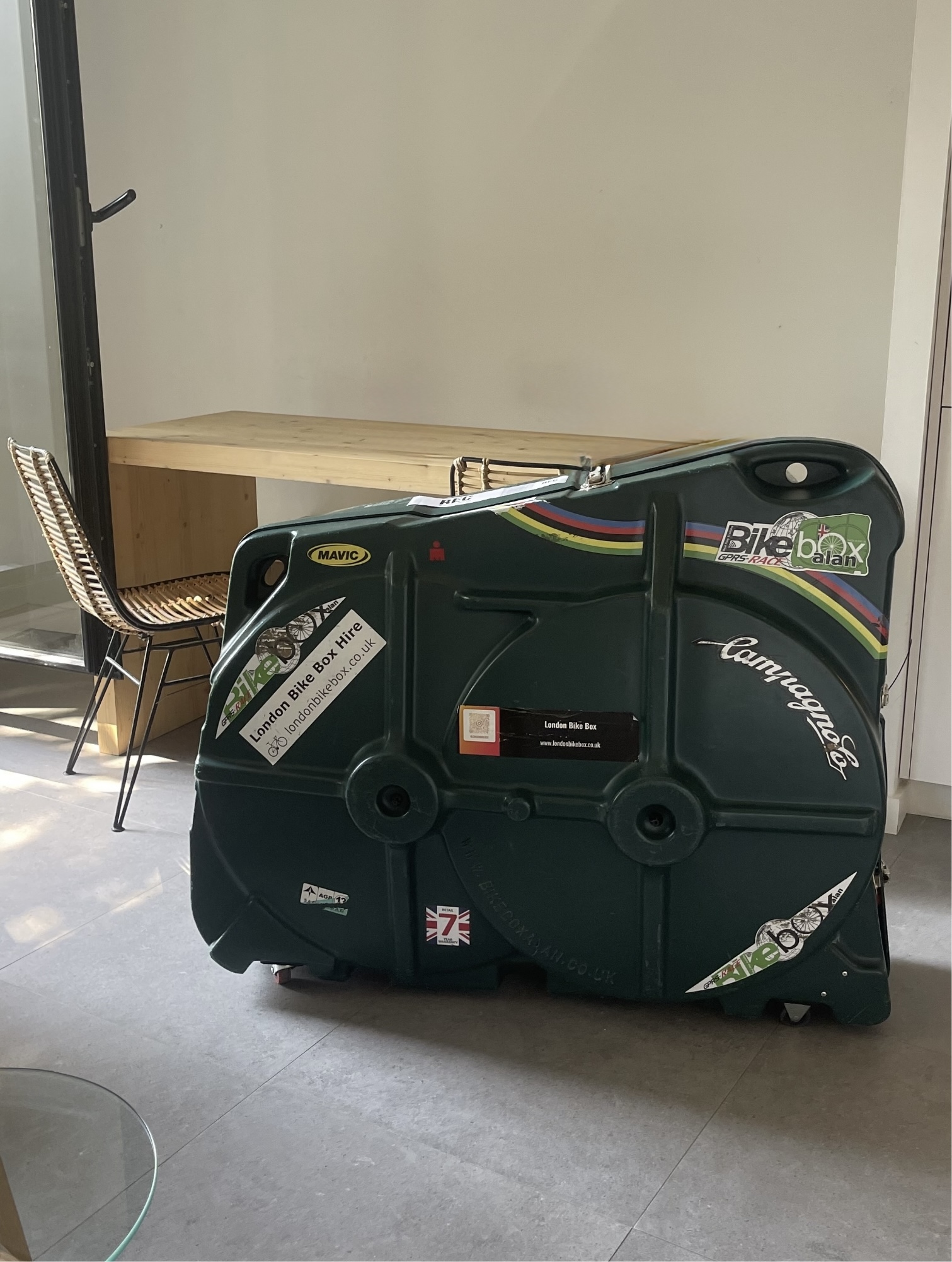 Large dark green hard bike travel case with colorful racing stripes, stickers including 'London Bike Box Hire' and 'Campagnolo', placed on a tiled floor in a room with a wooden table and wicker chair in the background.