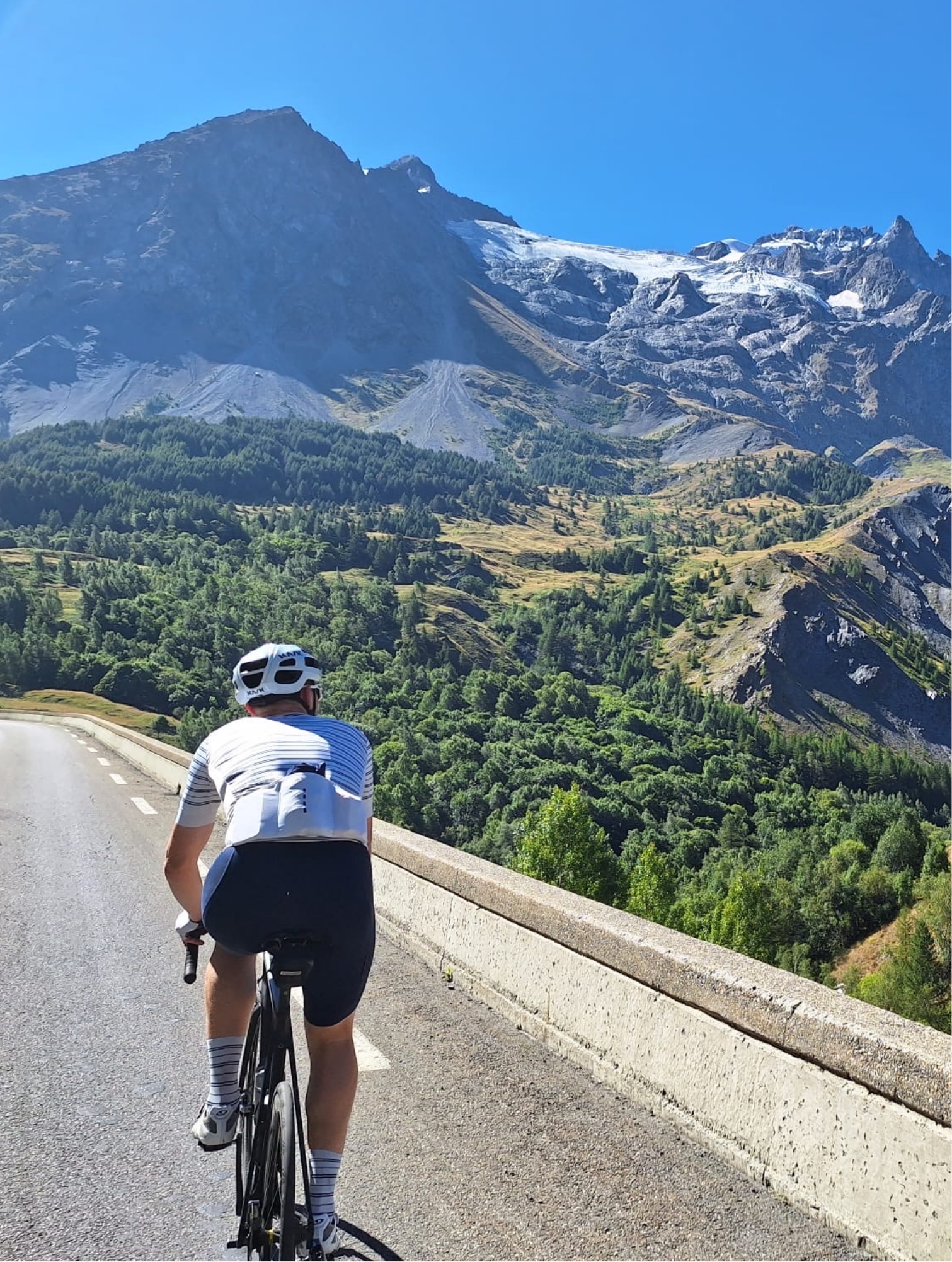 Person wearing a white helmet and cycling gear rides a road bike uphill on a mountain road with dense green trees below and large rocky peaks with patches of snow under a clear blue sky.