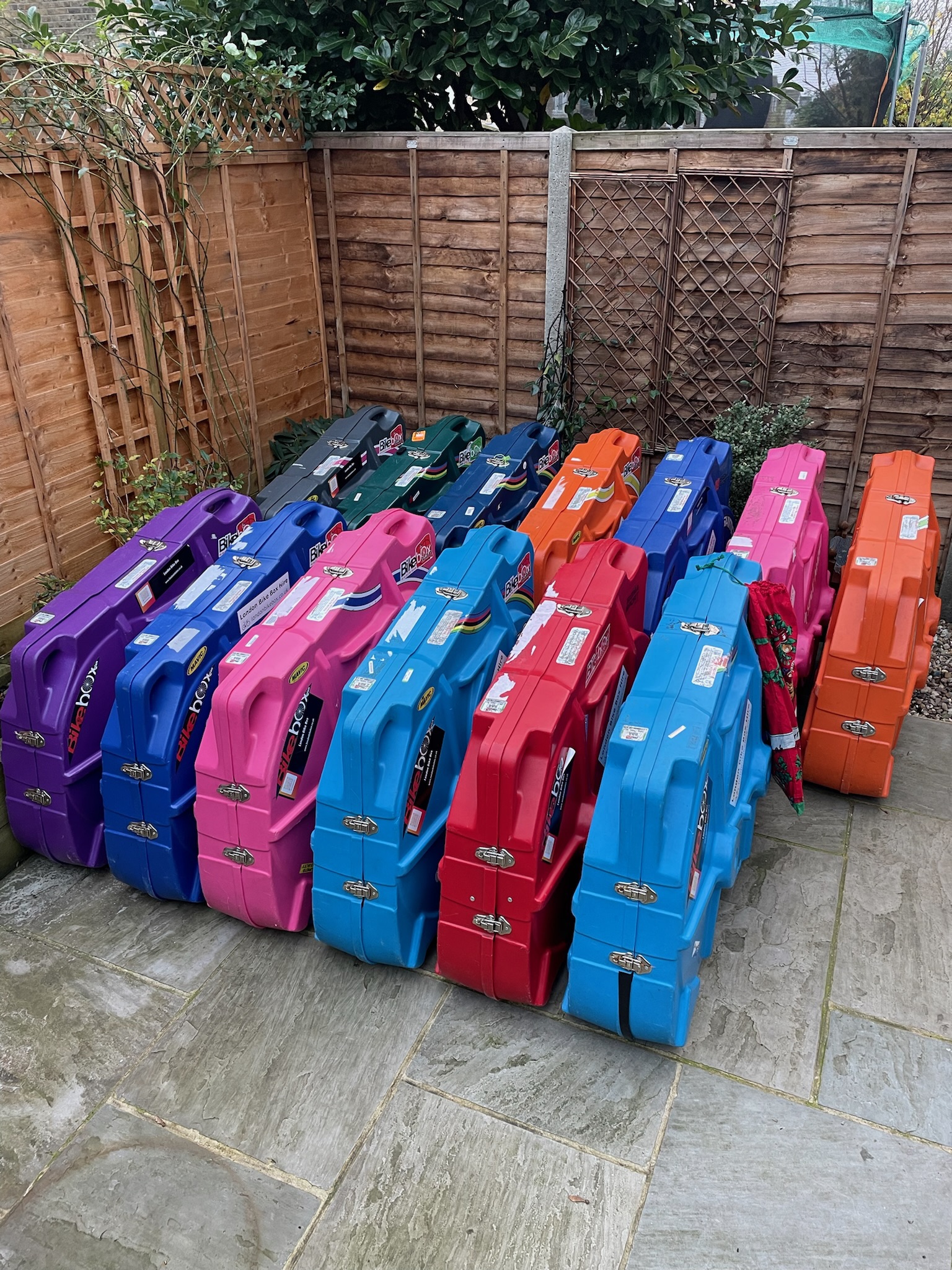 Fourteen colorful hard bike cases arranged in three rows on a stone patio next to wooden fences and greenery, with colors including purple, blue, pink, orange, and red, each with metal latches and labels.