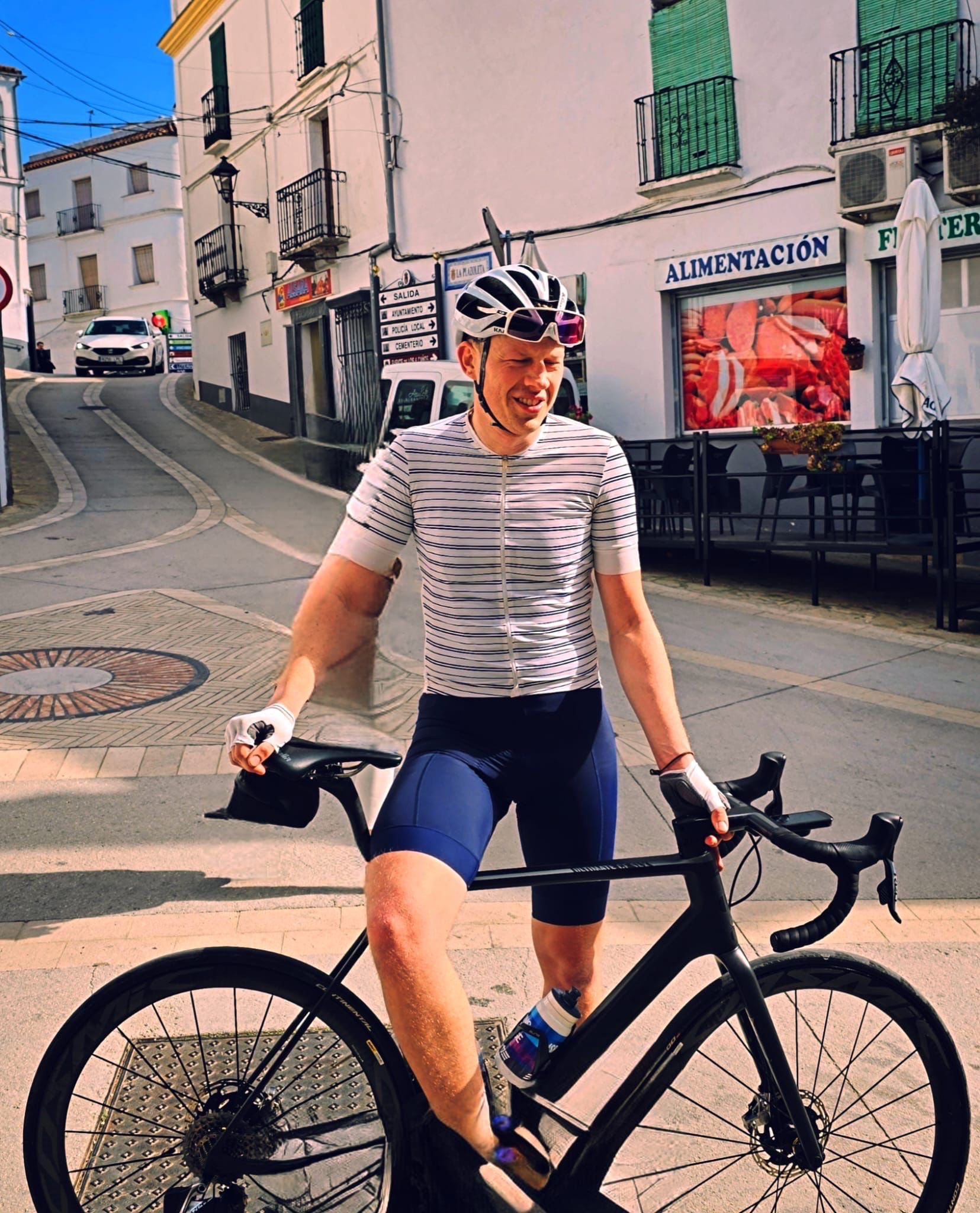 Male cyclist wearing a striped white and navy jersey, navy shorts, a white helmet, and sunglasses resting on a black road bike on a sunny street in a small town with white buildings and green window shutters.