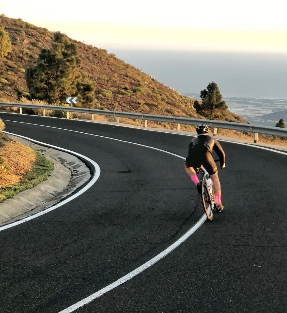 Cyclist wearing black gear and bright pink socks rides uphill on a winding mountain road with guardrails, surrounded by trees and vegetation, overlooking a distant coastal landscape under a clear sky.