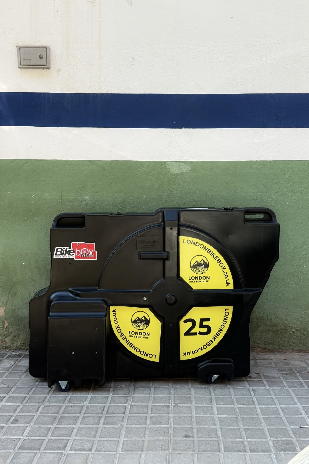 Black hard case bike box with yellow labels displaying the number 25 and the text 'London Bike Box Hire' with a bicycle and mountain logo, placed on a tiled floor against a green and white wall with a blue stripe.