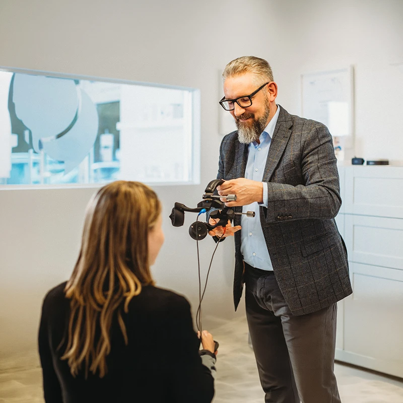 Ein Mann mit Brille und grauem Bart hält ein Audiometer, während eine Frau ihm gegenüber sitzt und das Gerät hält, in einem hellen Raum mit Fenster und Schränken.