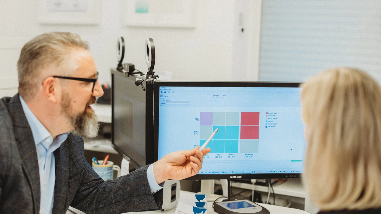 Ein Mann mit Brille und Bart erklärt einer Frau am Computerbildschirm eine farbige Diagrammtafel mit verschiedenen Lautstärkekategorien, die von extrem laut bis viel leiser reichen.