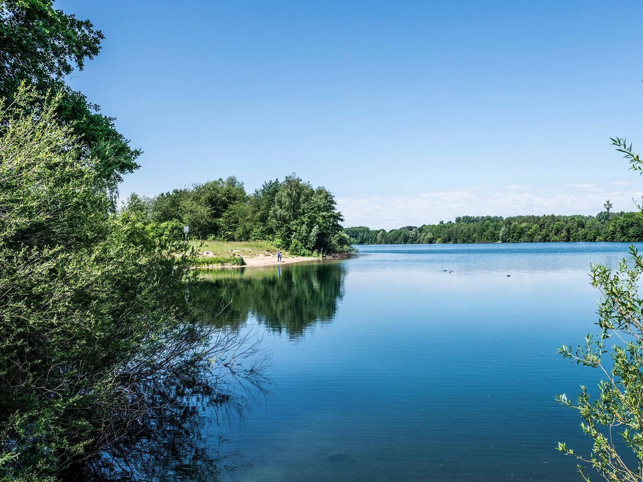 Blick auf einen ruhigen See mit klarem blauem Wasser, umgeben von grün bewachsenen Ufern unter einem hellblauen Himmel, mit zwei Menschen am linken Ufer und einem kleinen Turm am rechten Horizont.