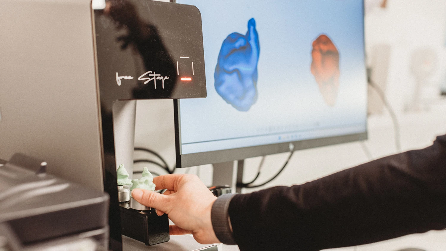 Eine Hand positioniert gedruckte Ohrstücke in einem 3D-Scanner, im Hintergrund zeigt ein Monitor blaue und rote 3D-Modelle von Ohren.