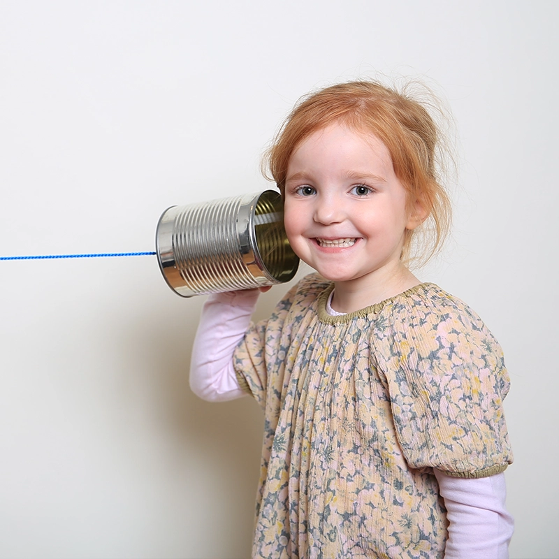Ein lachendes kleines Mädchen mit roten Haaren hält eine mit einer blauen Schnur verbundene Blechdose an ihr Ohr, die als improvisiertes Telefon dient.